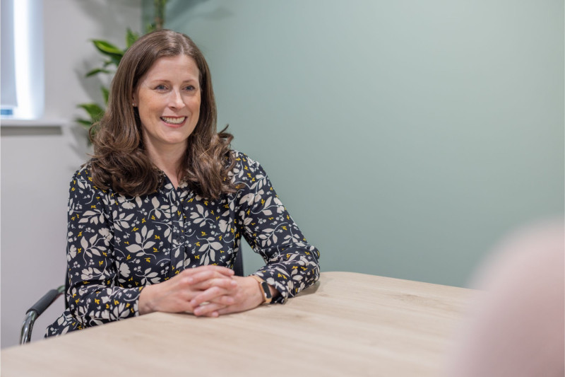 A woman with long brown hair smiles while seated at a table, wearing a patterned black blouse. She is engaged in conversation, with a plant visible in the background and a light green wall behind her.