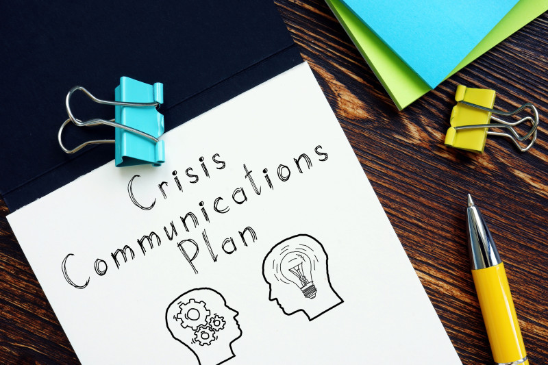 Crisis communications plan document with illustrations of gears and a lightbulb, accompanied by binder clips and a yellow pen on a wooden desk.