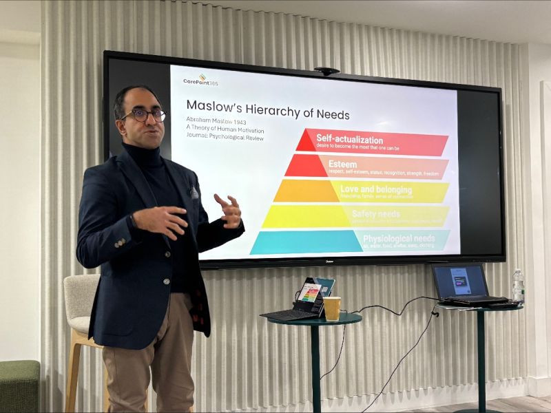 A speaker presents Maslow's Hierarchy of Needs on a screen during a conference, highlighting the five levels: Physiological needs, Safety needs, Love and belonging, Esteem, and Self-actualization.