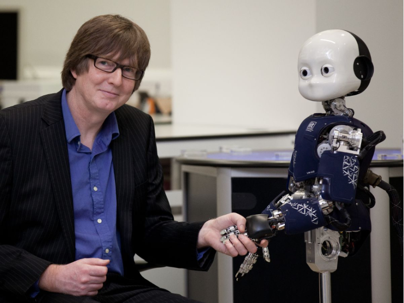 A man in a black suit and blue shirt sits beside a small humanoid robot, holding its hand. The robot has a rounded head and a blue torso with intricate patterns.